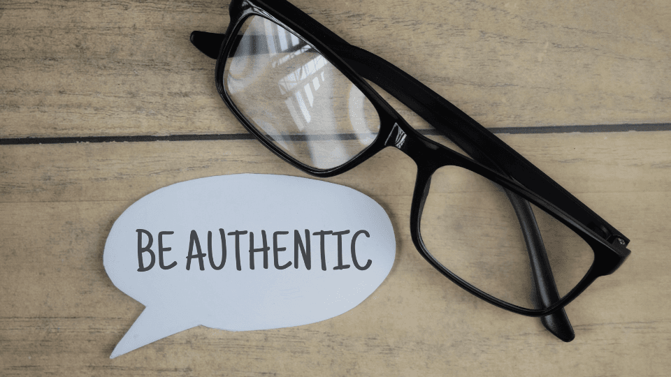 Image of a paper written "be authentic" next to it is a pair of glasses with a black frame.  The background is a wooden beige.