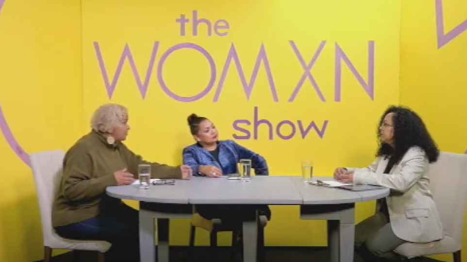 Three women sitting around a table having a conversation in a TV studio with a yellow background and The Womxn Show written on the wall. 