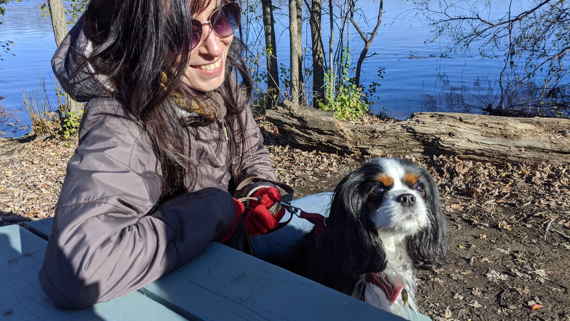 Woman with long brown hair wearing sunglasses sitting on a table outside next to a lake with her dog. She is smiling at the dog. 
