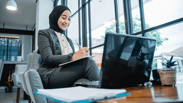 A confident young woman in a professional suit sits by the window in her Dubai office, smiling as she takes notes while working on her laptop, reflecting the spirit of determination and growth in the corporate world.