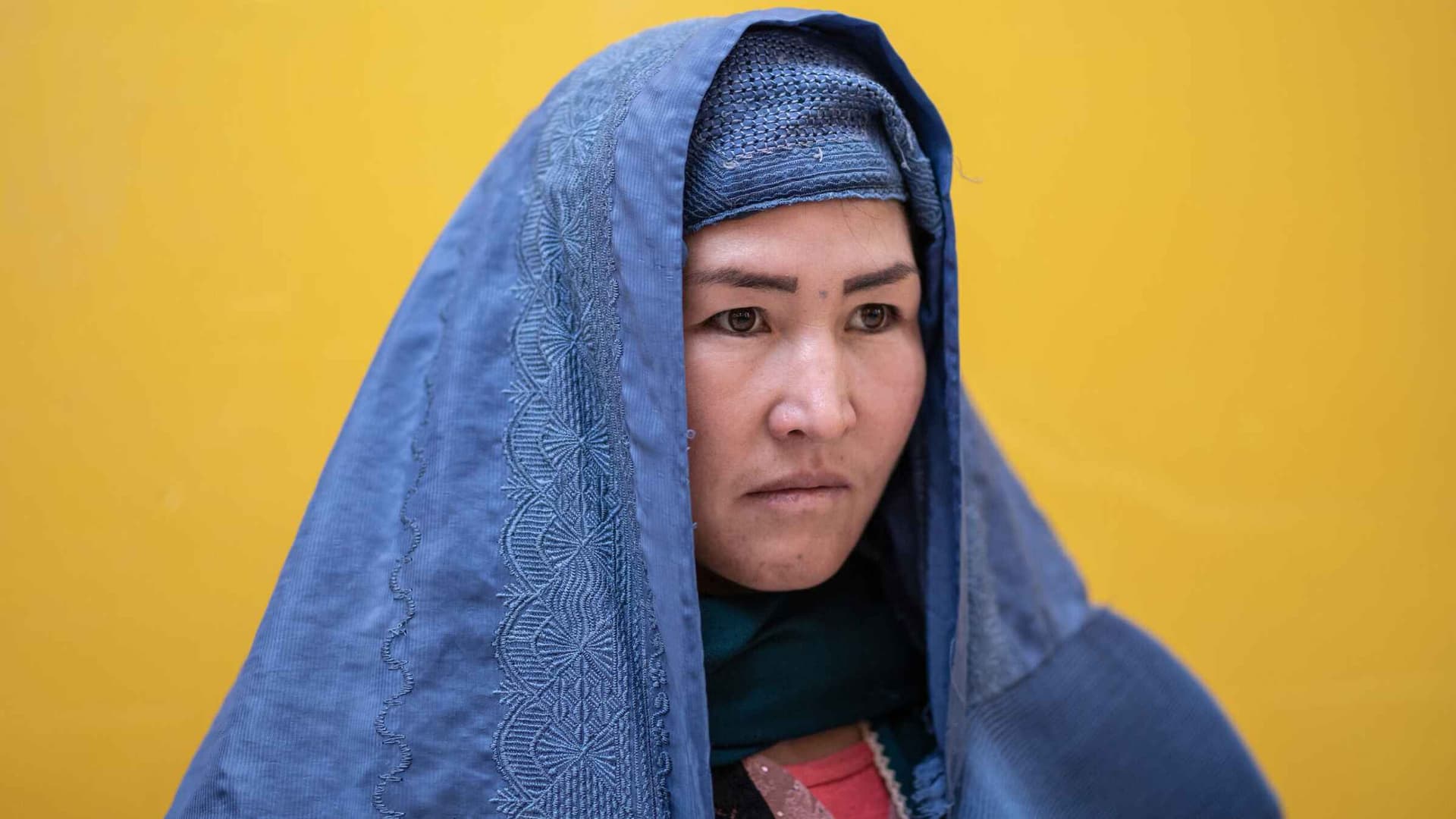 A woman in a blue hijab with a determined look, and a yellow wall in the background