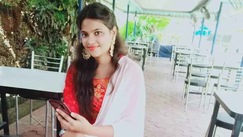  Seated on a patio with an overhang surrounded by tables and chairs, Sweta Singh sits holding a cell phone. She has long dark hair, gold earrings, a red dress, and white shawl.