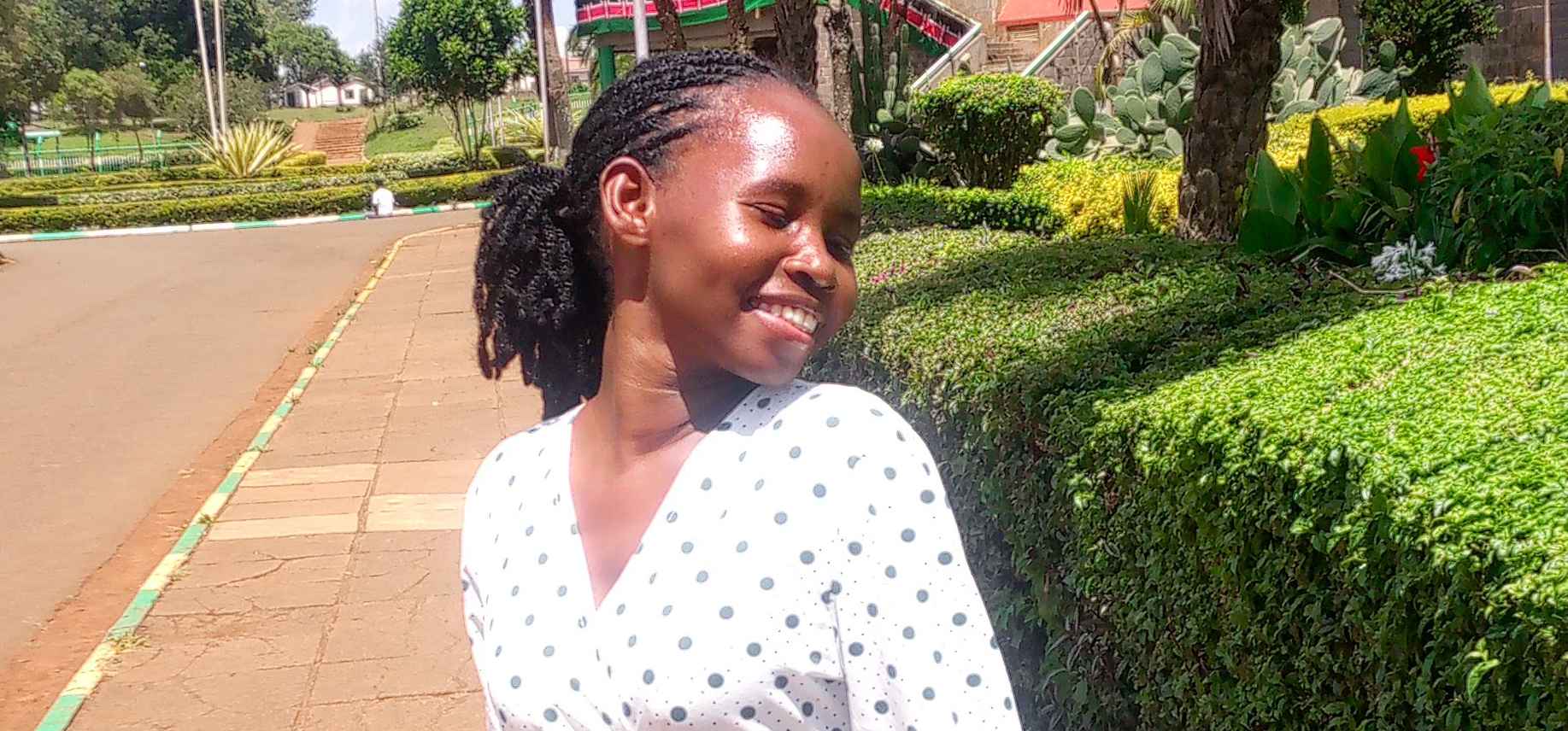 A smiling young woman wearing a white polka-dot dress stands outdoors, looking to her left. Green trees and a pathway create a peaceful natural background.